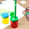 Two translucent pencil sharpeners (red with green lid and yellow with blue lid) on a wooden desk, a green pencil inserted into the red sharpener, and colored pencils nearby.